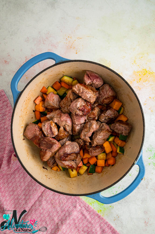 Browned lamb chunks added back to a Dutch oven with diced sweet potatoes, carrots, and zucchini.