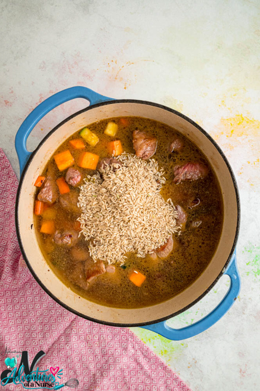 Uncooked brown rice added to a Dutch oven filled with lamb, vegetables, and broth.