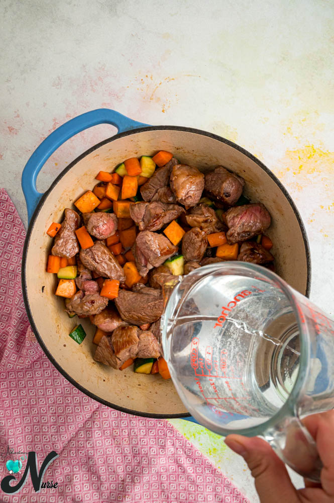 Water being poured from a glass measuring cup into a pot of lamb and vegetables.